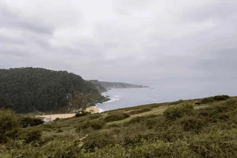 Paisaje costero con acantilados verdes y mar gris. Ideal para senderismo en Villaviciosa, con una playa dorada y una casa blanca.