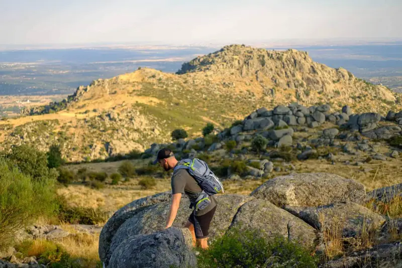 Un excursionista con mochila asciende por un terreno rocoso en la ruta Las Machotas, con un paisaje montañoso y un valle al fondo.